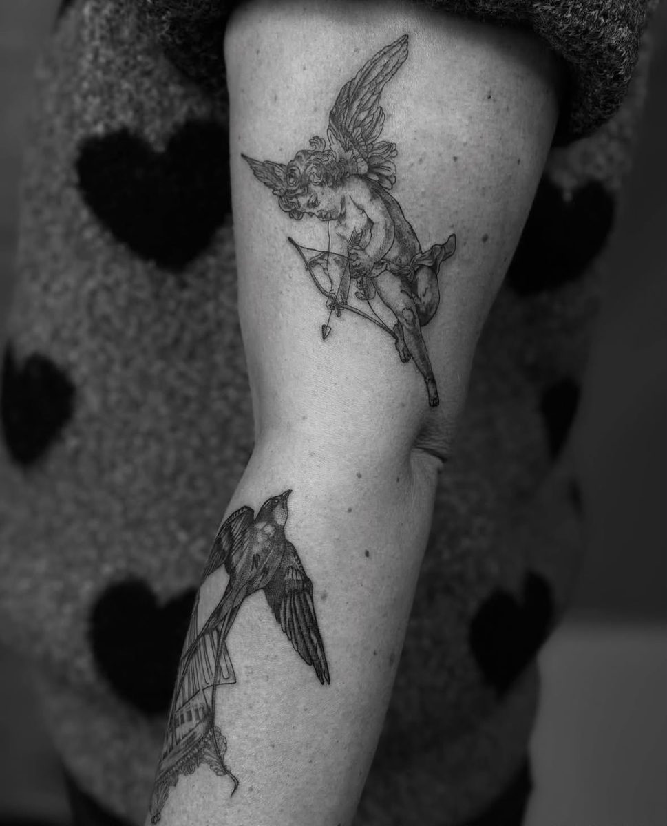 Tatouage Cupidon et hirondelle sur le bras par Nikita, NKT.INK